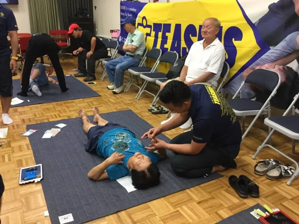 床に敷かれたマットの上で指導者が高齢者の手を触りながら説明する健康・介護予防体験会の光景。